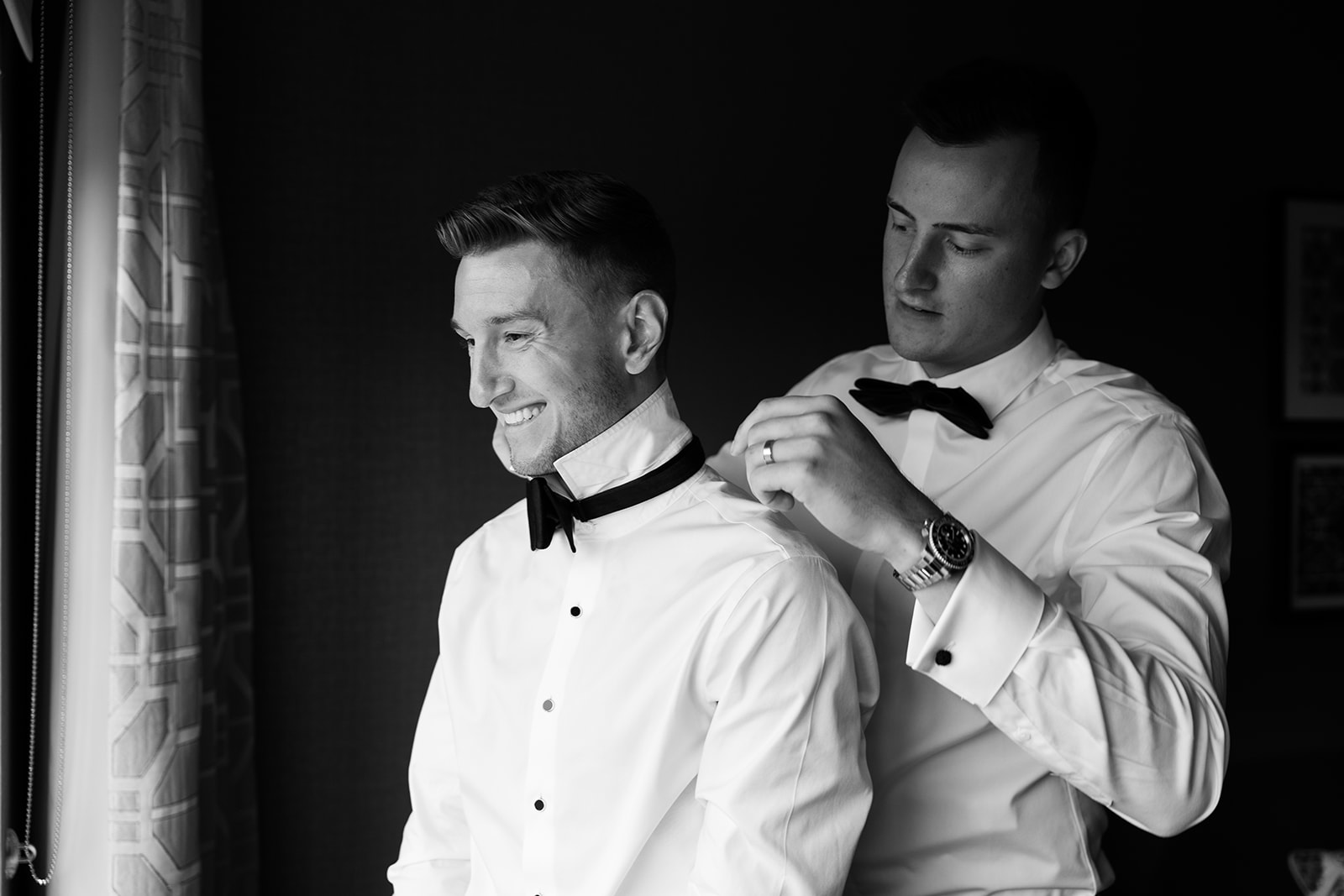 Black and white photo of a groom smiling as his groomsman adjusts his bow tie, sharing a warm moment.